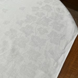 May include: White tablecloth with a delicate leaf pattern. The fabric has a textured finish, and the design features repeating ivy leaves. The tablecloth is draped over a round surface, showing the edge and corner.