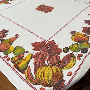 May include: A white square tablecloth featuring a decorative border of colourful autumn fruits including pumpkins, pears, apples, and grapes, accented with brown leaves. A matching cluster of leaves is centred in the middle of the fabric.