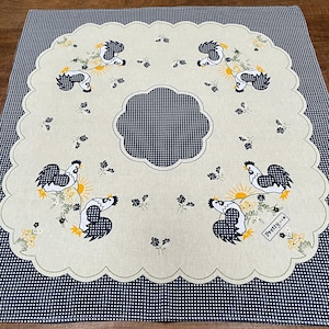 May include: A square fabric tablecloth featuring a dark blue and white gingham border and a central scalloped cream panel. The cream section displays a repeating pattern of roosters with dark blue checkered wings, yellow suns, and small flowers.