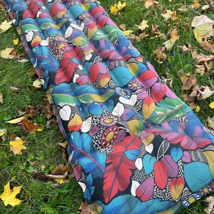Peut inclure: Un coussin long et rectangulaire, à motifs noirs et bleus, avec une surface texturée. Le coussin présente un motif floral coloré avec des accents rouges, jaunes, verts et blancs. Le coussin est posé sur un lit d'herbe verte avec des feuilles d'automne tombées.