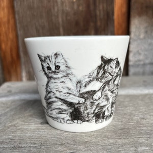 Peut inclure: Une tasse en céramique blanche avec une illustration en noir et blanc de deux chatons qui jouent. Les chatons sont dessinés dans un style réaliste et ont une expression espiègle.