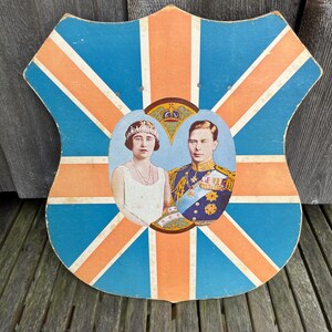 Peut inclure: Une plaque murale en forme de bouclier arborant un motif Union Jack bleu et orange. Au centre, un portrait ovale représente une femme en robe blanche avec un diadème et un homme en uniforme militaire décoré, surmonté d'un emblème de couronne.