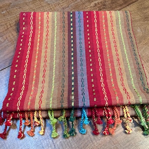 Peut inclure: Textile rectangulaire plié, à base rouge et rayures verticales multicolores, dont vert, jaune et bleu. Les bords sont ornés de pompons colorés. Le textile semble être tissé.