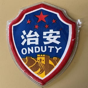 Peut inclure: Écusson en forme de bouclier avec un fond bleu, une bordure rouge et une bordure blanche. L'écusson comporte des caractères chinois, le texte anglais "ONDUTY", une étoile rouge et de petites étoiles rouges. Une illustration marron et jaune d'un bâtiment est en bas.