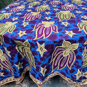 May include: A colourful tablecloth featuring a repeating floral and star design. The flowers are purple, green, and gold, with white detailing. Gold stars and a gold border complete the design.