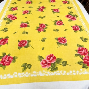 May include: A rectangular tablecloth featuring a vibrant yellow background with a repeating pattern of pink roses and green leaves. The design includes a white decorative border with a scalloped edge pattern framing the floral print.