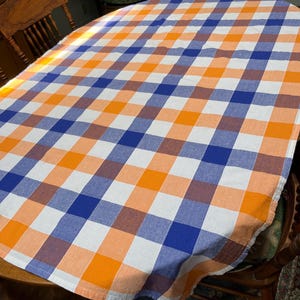 May include: An oval tablecloth featuring a large-scale plaid pattern in vibrant orange, light peach, deep blue, and white. The fabric displays a woven texture with a consistent grid design that covers the entire surface of the oval-shaped textile.