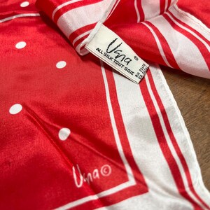 May include: A red and white silk scarf with a polka dot and striped pattern. The scarf has a tag that reads "Una" and "All Silk Tout Soie". The scarf is folded, showing the different patterns.