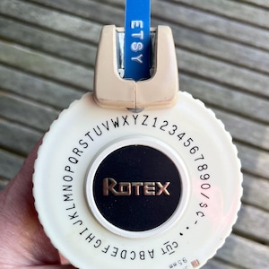 Peut inclure: Une étiqueteuse Rotex vintage, crème et noire avec un centre doré. Une étiquette bleue affiche "ETSY". Le cadran présente des lettres, des chiffres et des symboles. La largeur de coupe est de 9,5 mm.