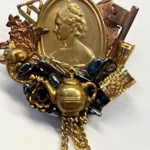 Peut inclure: Une broche dorée avec un camée représentant le profil d'une femme, entouré de divers éléments décoratifs, dont une théière, une clé et un dé à coudre. La broche a une chaîne pendante.