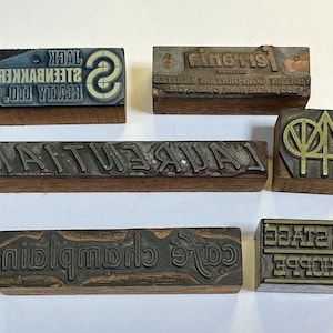 May include: Six wooden printing blocks with various text and designs. The blocks are all different sizes and shapes. The text on the blocks includes "REALTY LTD", "STEENBAKERS", "JACK", "S", "FERRANIA", "LAUREL AUTUMN", "SHOPPE", "YE STAGE", and a stylised tree design.