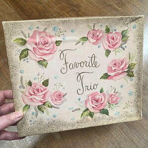 May include: A vintage box with a light brown background and a gold glitter texture. The box is decorated with pink roses and blue flowers. The text "Favorite Trio" is written in black cursive.