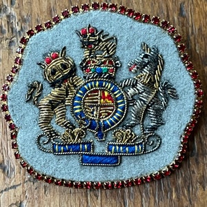 May include: A gold embroidered patch featuring a royal coat of arms with a lion, a unicorn, and a crown. The patch is surrounded by a border of red rhinestones.