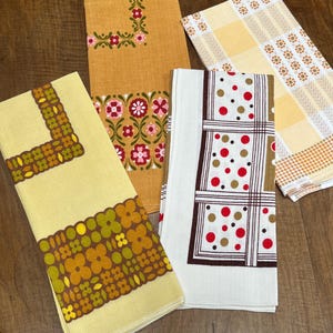May include: A selection of vintage kitchen towels with diverse patterns and colours. The towels display floral and geometric designs in yellow, orange, brown, and white. Some have decorative borders and polka dots.