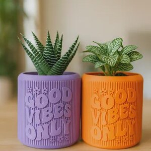 May include: A purple and an orange cylindrical planter, each featuring vertical ribbed textures and the embossed text "GOOD VIBES ONLY" with small flower accents. The purple planter holds a green succulent, while the orange planter holds a green plant.
