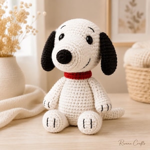 Puede incluir: Una figura de perro tejida a ganchillo en blanco y negro con cabeza redonda, orejas negras caídas, nariz negra y collar rojo. El personaje tiene ojos y sonrisa bordados en negro, con pequeñas líneas negras en las patas. El texto Rivana Crafts es visible.