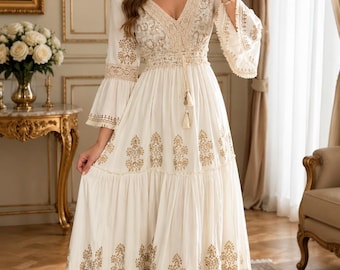 May include: A floor-length cream-colored dress featuring a deep V-neckline, bell sleeves, and a cinched waist with a drawstring tassel tie. The garment is adorned with intricate gold-toned embroidery and horizontal bands of beige lace trim.