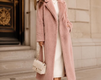 May include: A long, dusty pink faux fur coat with a wide lapel collar and long sleeves, worn over a cream-colored ribbed turtleneck dress. The outfit is paired with cream-colored ankle boots and a cream-colored quilted shoulder bag with a gold chain.