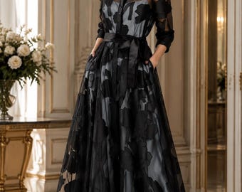 May include: A floor-length black shirt dress featuring a sheer overlay with a large floral pattern, a collared neckline, long sleeves, and a solid black waist tie. The dress has a full skirt with side pockets and a light gray inner lining.