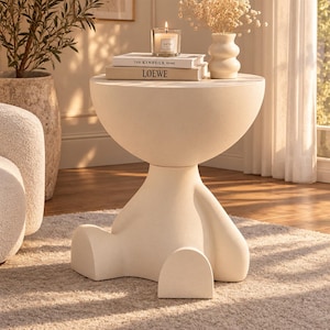 Puede incluir: Una mesa auxiliar de color crema con una superficie lisa y redondeada y una base con forma de figura sentada con dos pies salientes. La mesa sostiene una vela encendida en un frasco de vidrio y dos libros, The Kinfolk Home y LOEWE.