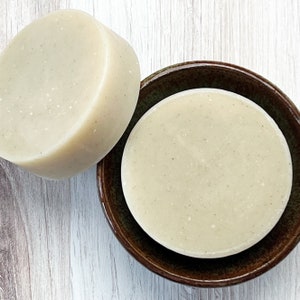 May include: Two round, off-white soap bars. One bar is on a brown ceramic dish, and the other is beside it.