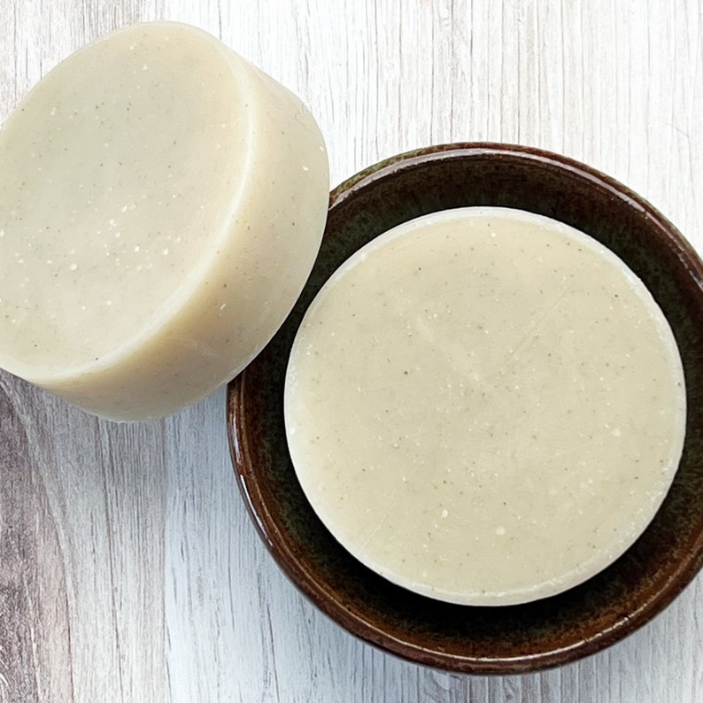 May include: Two round, off-white soap bars. One bar is on a brown ceramic dish, and the other is beside it.