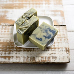 May include: Three rectangular bars of handmade soap with a marbled design in shades of green and blue, presented on a white square plate. The soap has a light green base color with darker green and blue swirls. The soap is displayed on a rustic wooden surface.