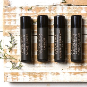 May include: Four black lotion sticks with white text, each labeled with a different scent: Balsam + Juniper, Lavender, Grapefruit Bergamot, and Ylang Ylang Lime. The sticks are on a distressed wooden surface with sprigs of greenery.