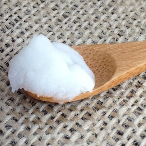 May include: A small wooden spoon filled with a white, creamy substance, likely coconut oil or butter, on a woven burlap background.