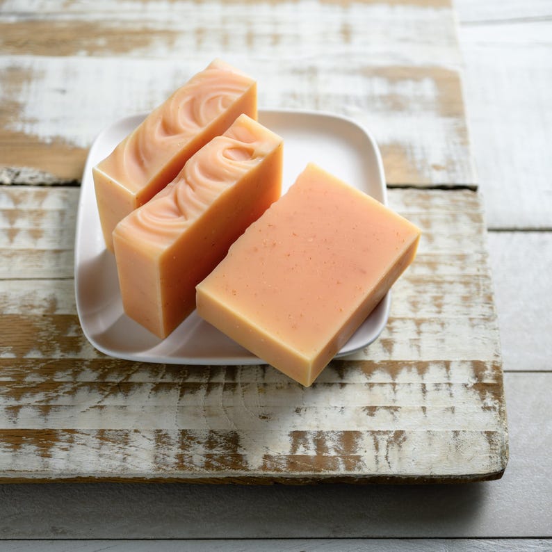 May include: Four bars of peach-colored soap on a white square plate. The soap has a smooth, rectangular shape with a swirl design on the top of three bars. The plate sits on a distressed, white-painted wooden surface.