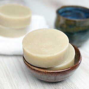 May include: Two round bars of natural soap in a brown ceramic dish. The soap is a light beige color and has a speckled appearance.