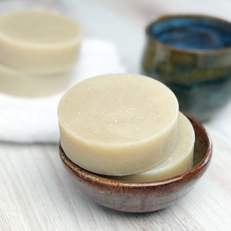 May include: Two round bars of natural soap in a brown ceramic dish. The soap is a light beige color and has a speckled appearance.