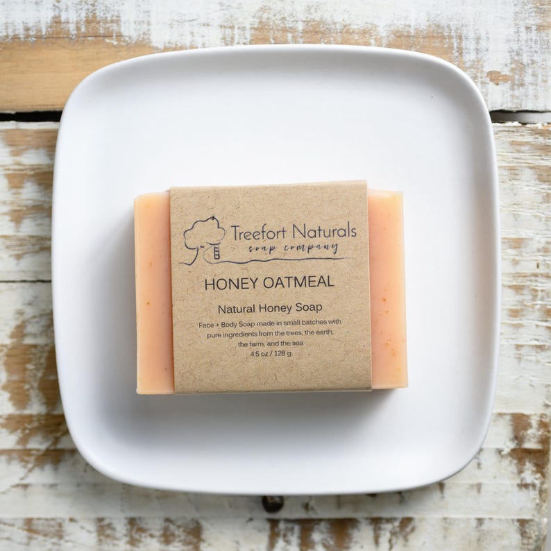 May include: A bar of Honey Oatmeal natural soap from Treefort Naturals, resting on a white square plate. The soap is light orange and wrapped in a brown paper label. The label reads "HONEY OATMEAL" and "4.5 oz / 128 g."
