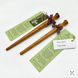 Puede incluir: Dos utensilios de cocina spurtle de madera marrón claro con mangos cónicos y extremos redondeados. Cada spurtle está atado con una cinta de cuadros roja, verde y azul. Las etiquetas adjuntas muestran el texto Turning Wood Works, Cherry Wood y Prunus avium.