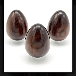 May include: Three smooth, polished, egg-shaped objects crafted from dark brown Santos rosewood with visible swirling wood grain patterns. Each egg is displayed against a plain white background, showcasing a glossy finish and natural wood texture.
