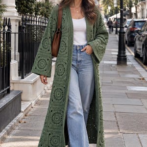 May include: An olive green floor-length crochet cardigan featuring a repeating square motif with circular patterns, worn over a white tank top and light blue denim jeans, paired with a brown shoulder bag and white sneakers.