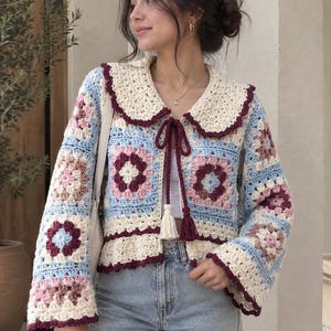 May include: A long-sleeved crochet cardigan featuring a granny square pattern in shades of light blue, cream, and dusty pink. The garment includes a large cream-colored rounded collar with a dark burgundy trim and a front tie closure with tassels.