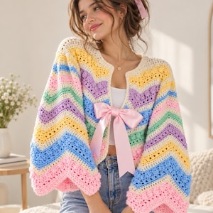 May include: A long-sleeved crochet cardigan featuring a chevron pattern in pastel shades of yellow, purple, blue, green, and pink. The garment has a wide, open front design secured by a large, tied light pink satin ribbon bow at the center.