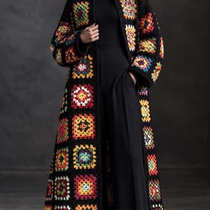 May include: A long, open-front crochet cardigan featuring a black base with multicolored granny square motifs in shades of red, yellow, blue, green, and purple. The garment has long sleeves and a floor-length hem, worn over a black turtleneck and trousers.