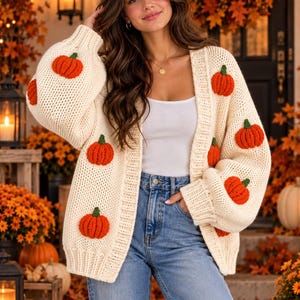 May include: A cream-colored chunky knit cardigan featuring a repeating pattern of small, raised orange pumpkins with green stems. The open-front sweater has long, voluminous sleeves, a ribbed collar, and ribbed cuffs, worn over a white tank top.