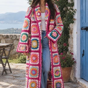 May include: A long, open-front cardigan made from multicolored granny square crochet patches. The garment features a vibrant fuchsia border and a repeating pattern of squares in shades of yellow, light blue, pink, and white, with long sleeves.