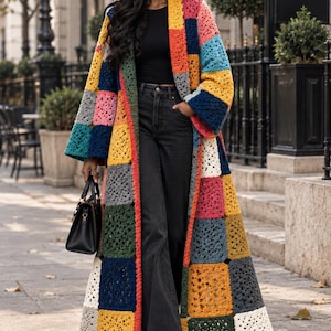 May include: A long, open-front cardigan made of multicoloured crochet granny squares in shades of yellow, blue, pink, green, grey, and white. The garment features long sleeves and a straight silhouette, worn over a black top and black jeans.