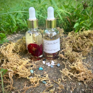 Puede incluir: Dos frascos cuentagotas de vidrio transparente con cuellos dorados y bulbos de goma blancos. Un frasco contiene un pétalo de rosa roja, mientras que el otro tiene una etiqueta blanca con el texto escrito a mano Custom Ritual Oil - Self-love, healing.