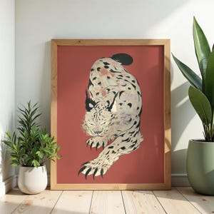 Peut inclure: Une impression d'art encadrée représentant une illustration stylisée d'un lynx en position de bond. Le lynx a un pelage crème avec des taches noires et des touffes d'oreilles sombres, sur un fond rouge terre cuite dans un cadre en bois clair.