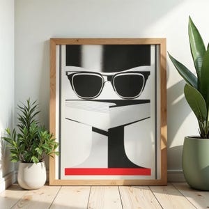 Peut inclure: Une impression d'art graphique encadrée présentant une illustration stylisée en noir et blanc d'une paire de lunettes à monture sombre posée sur un livre, avec une bande horizontale rouge vive en bas, le tout dans un cadre en bois clair.