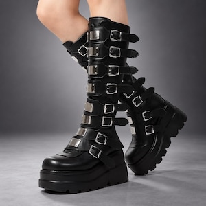 Könnte beinhalten: Schwarze wadenhohe Stiefel mit einer dicken Plateausohle mit Profil. Die Stiefel sind mit mehreren horizontalen Riemen verziert, die jeweils mit einer silberfarbenen Metallschnalle befestigt und vorne mit einer rechteckigen silbernen Metallplatte versehen sind.