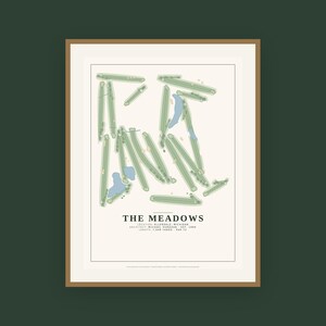 Op de afbeelding: Een ingelijste print met een plattegrond van een golfbaan genaamd THE MEADOWS, gelegen in Allendale, Michigan. Het ontwerp bevat groene fairways, blauwe waterhindernissen en tekst met details over de architect, het oprichtingsjaar en de lengte.