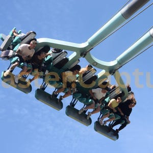 May include: A suspended roller coaster train with teal metal supports and individual seats carrying passengers high against a clear blue sky. The text Coaster.Capture is overlaid across the centre of the image in a light, semi-transparent font.