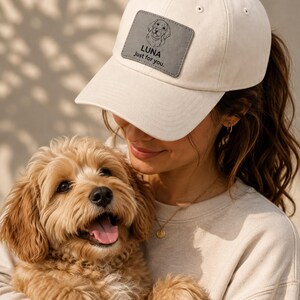 Può includere: Un cappellino da baseball color crema con una toppa rettangolare grigia sul davanti. La toppa mostra un'illustrazione a linee del muso di un cane sopra il testo LUNA e la frase Just for you. Il cappellino ha una corona strutturata e visiera.