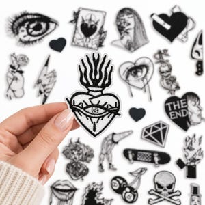 May include: A black and white embroidered patch featuring a sacred heart design with flames on top and an eye in the center. The patch is held against a background of various other black and white embroidered patches with diverse designs.